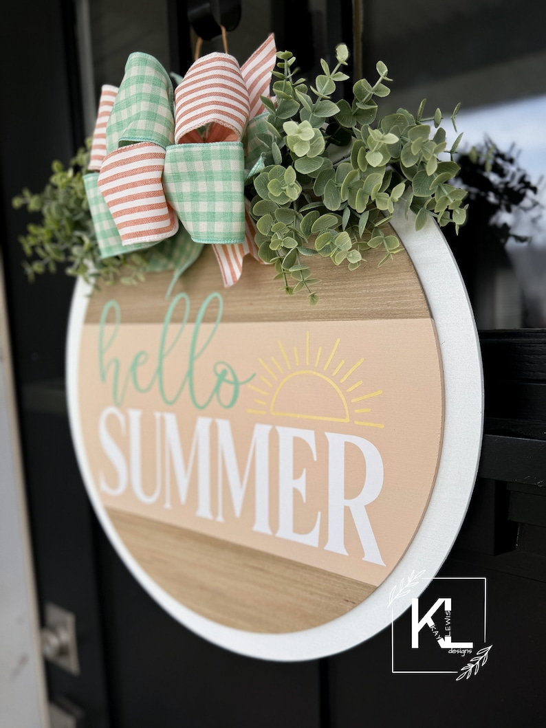 Pu&ograve; includere: Un appendiporta rotondo in legno con un bordo bianco. L'appendiporta ha uno sfondo pesca con un sole giallo e le parole "hello summer" in lettere bianche e verdi. Un fiocco di Vichy verde e bianco e un ramoscello di verde sono attaccati alla parte superiore dell'appendiporta.
