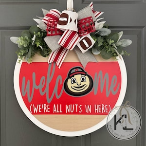 Puede incluir: Un letrero de puerta redondo de madera con acabado en rojo y madera natural. Incluye el texto "welcome" en gris y "(WE'RE ALL NUTS IN HERE)" en blanco. Una cabeza de mascota de dibujos animados reemplaza la "o". Un lazo con patrones de balones de f&uacute;tbol y rayas corona el dise&ntilde;o junto a follaje verde.