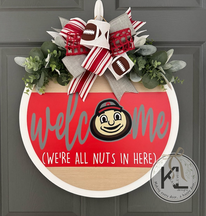 Puede incluir: Un letrero de puerta redondo de madera con acabado en rojo y madera natural. Incluye el texto "welcome" en gris y "(WE'RE ALL NUTS IN HERE)" en blanco. Una cabeza de mascota de dibujos animados reemplaza la "o". Un lazo con patrones de balones de f&uacute;tbol y rayas corona el dise&ntilde;o junto a follaje verde.