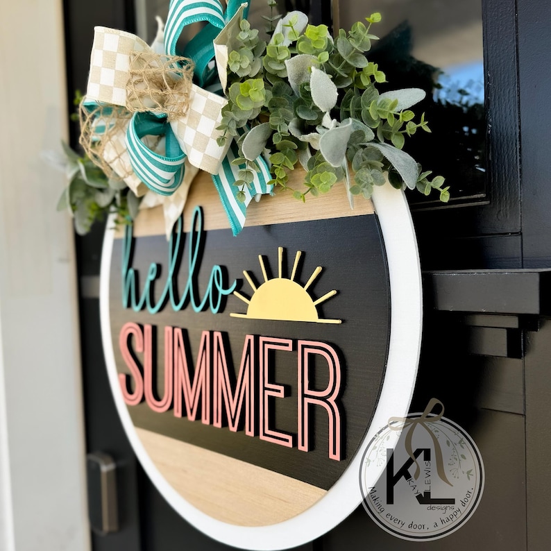 May include: A round wooden door hanger with a white border. The sign says "hello summer" in colorful letters with a yellow sun graphic. The sign is decorated with a green and white bow.