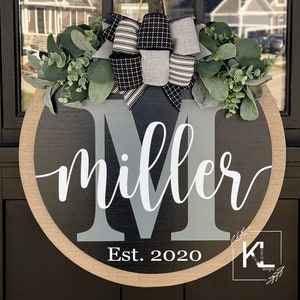 Puede incluir: Un colgador de puerta redondo de madera negro y gris con una gran letra M gris y el nombre "miller" escrito en cursiva blanca. El a&ntilde;o "Est. 2020" est&aacute; escrito debajo del nombre. El colgador de puerta est&aacute; decorado con un lazo a rayas blanco y negro y follaje verde.
