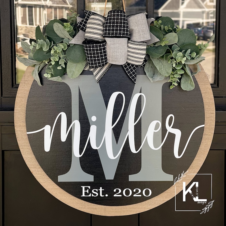 Puede incluir: Un colgador de puerta redondo de madera negro y gris con una gran letra M gris y el nombre "miller" escrito en cursiva blanca. El a&ntilde;o "Est. 2020" est&aacute; escrito debajo del nombre. El colgador de puerta est&aacute; decorado con un lazo a rayas blanco y negro y follaje verde.