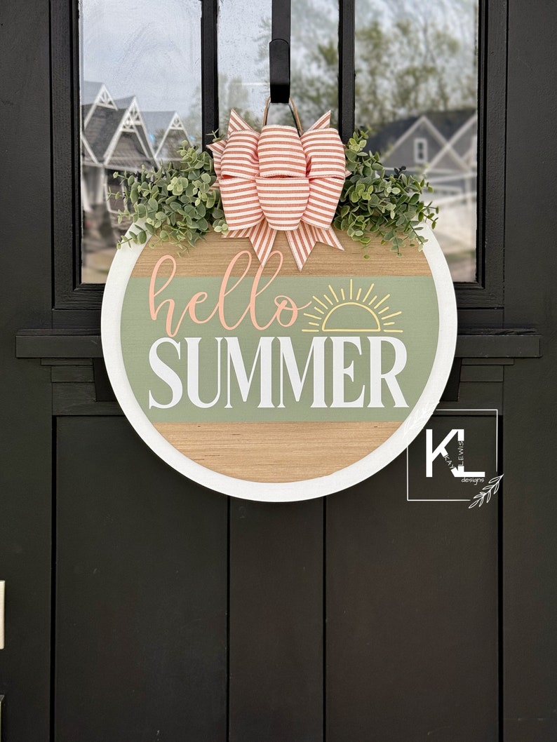Pu&ograve; includere: Un appendiporta rotondo in legno con un bordo bianco e uno sfondo verde e legno. Le parole "hello summer" sono scritte in bianco con una grafica di sole gialla. Un fiocco a righe rosa e bianco con del verde &egrave; attaccato alla parte superiore.