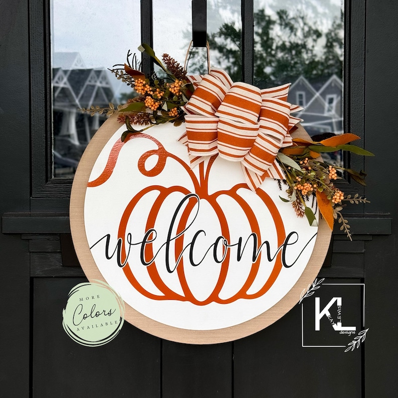May include: A round wooden door hanger with a white pumpkin design and the word "welcome" in black script. The pumpkin is outlined in orange and has a brown stem. A white and orange striped bow with fall foliage is attached to the top of the pumpkin.