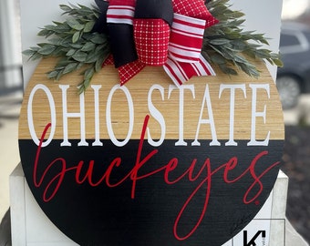 May include: A round wooden door hanger with a black and natural wood finish. The top half of the hanger is natural wood with the words "Ohio State" in white lettering. The bottom half is black with the word "buckeyes" in red cursive lettering. A red, white, and black bow with greenery is attached to the top of the hanger.