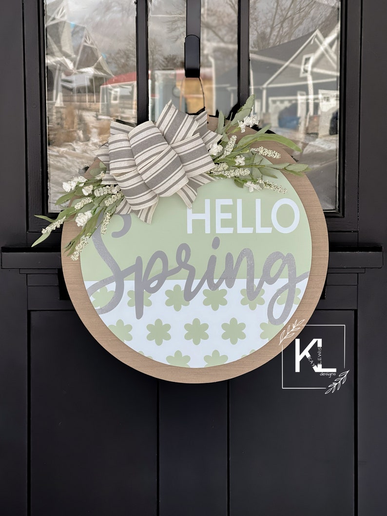 May include: A round wooden door hanger with a green and white floral pattern and the text "HELLO Spring" in white lettering. A gray and white striped bow is attached to the top of the hanger.