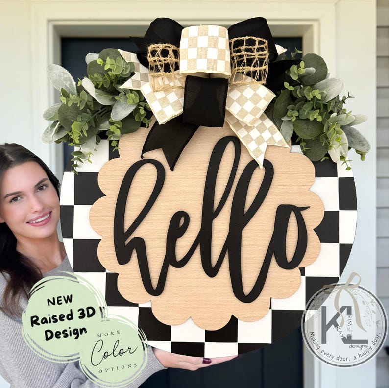 Op de afbeelding: Een ronde, zwart-wit geblokte houten deurhanger met het woord "hello" in zwart schrift. Het bord heeft een geschulpte rand en is versierd met een zwart-beige geblokte strik, groen en een zwarte strik. Het bord heeft ook de tekst "NEW Raised 3D Design" en "MORE Color OPTIONS".