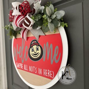 Puede incluir: Un letrero de puerta redondo de madera con un centro rojo que incluye el texto welcome y we're all nuts in here, una mascota de dibujos animados y un gran lazo decorativo con patrones de f&uacute;tbol americano, adornado con follaje verde artificial.