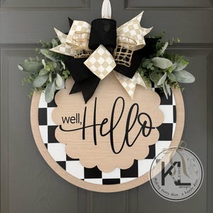 K&ouml;nnte beinhalten: Ein rundes T&uuml;rschild aus Holz mit schwarz-wei&szlig;em Karomuster, einer gewellten Mittelplatte mit dem Text "well, Hello" in schwarzer Schrift und einer dekorativen Schleife aus schwarz-wei&szlig;-beigem Karoband mit gr&uuml;nem Blattwerk.
