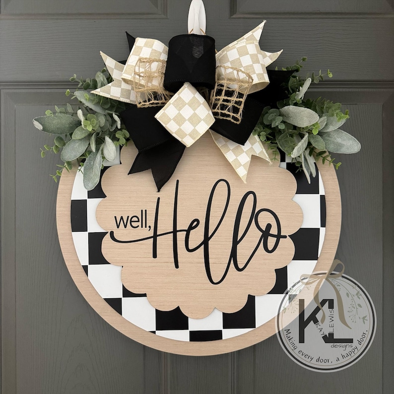 K&ouml;nnte beinhalten: Ein rundes T&uuml;rschild aus Holz mit schwarz-wei&szlig;em Karomuster, einer gewellten Mittelplatte mit dem Text "well, Hello" in schwarzer Schrift und einer dekorativen Schleife aus schwarz-wei&szlig;-beigem Karoband mit gr&uuml;nem Blattwerk.
