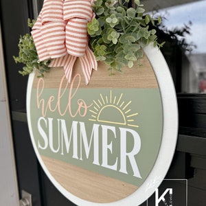 Pu&ograve; includere: Un cartello rotondo in legno con un bordo bianco, che presenta il testo "hello summer" in lettere rosa e bianche. Un sole stilizzato con raggi gialli &egrave; sopra il testo. Il cartello &egrave; decorato con un fiocco rosa e bianco a righe e foglie di eucalipto verdi.