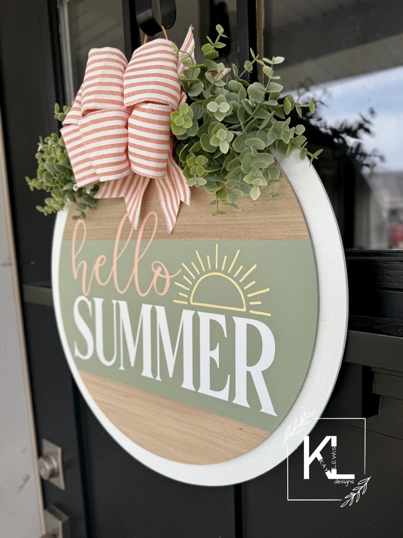 Pu&ograve; includere: Un cartello rotondo in legno con un bordo bianco, che presenta il testo "hello summer" in lettere rosa e bianche. Un sole stilizzato con raggi gialli &egrave; sopra il testo. Il cartello &egrave; decorato con un fiocco rosa e bianco a righe e foglie di eucalipto verdi.