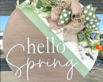 May include: A round wooden door hanger with a green and white striped design. The words "hello spring" are written in white script on the hanger. A green and brown bow with white polka dots is attached to the top of the hanger.
