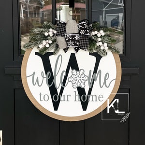 May include: A round wooden door hanger with a white background and a black letter "W" in the center. The words "welcome to our home" are written in gray cursive script around the letter. A white snowflake is centered on the letter "W". The door hanger is decorated with a white and black plaid bow and a sprig of faux evergreen with white berries.