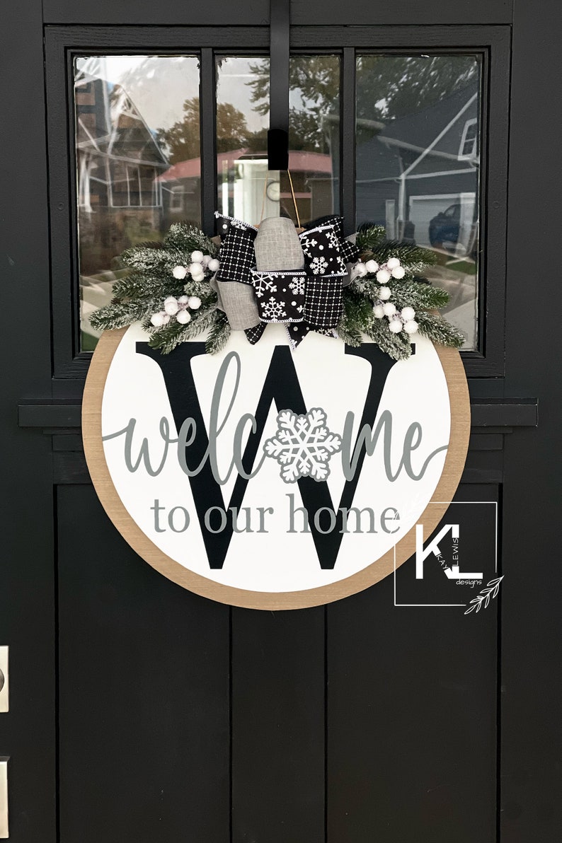 May include: A round wooden door hanger with a white background and a black letter "W" in the center. The words "welcome to our home" are written in gray cursive script around the letter. A white snowflake is centered on the letter "W". The door hanger is decorated with a white and black plaid bow and a sprig of faux evergreen with white berries.