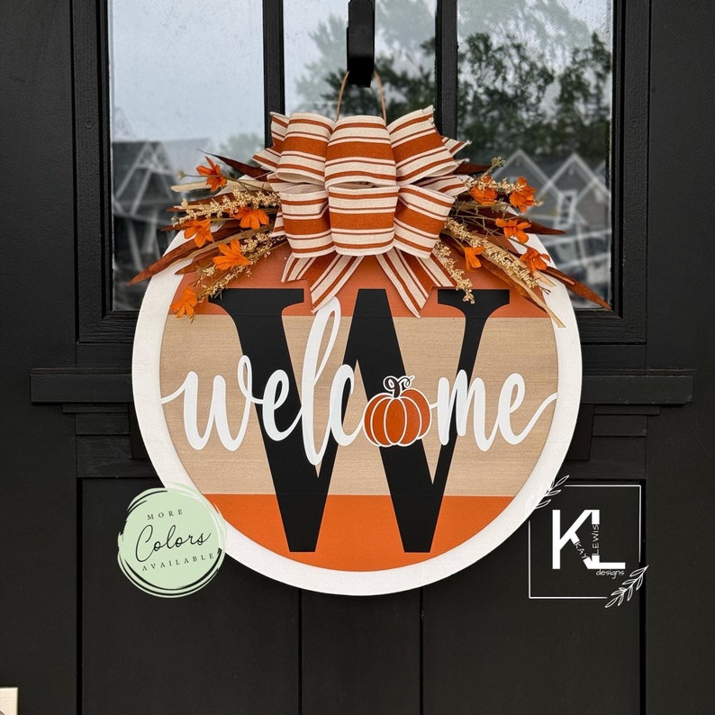 May include: A round wooden welcome sign with a black letter W and the word 'welcome' in white cursive. The sign has orange and brown stripes and a small orange pumpkin in the center of the W. The sign is decorated with a brown and white striped bow and orange and brown fall foliage. The sign has a sticker that says 'More Colors Available'.