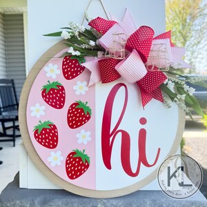 May include: A pink and white round door hanger with a red "hi" and a strawberry pattern. The door hanger is decorated with a pink and red polka dot bow.