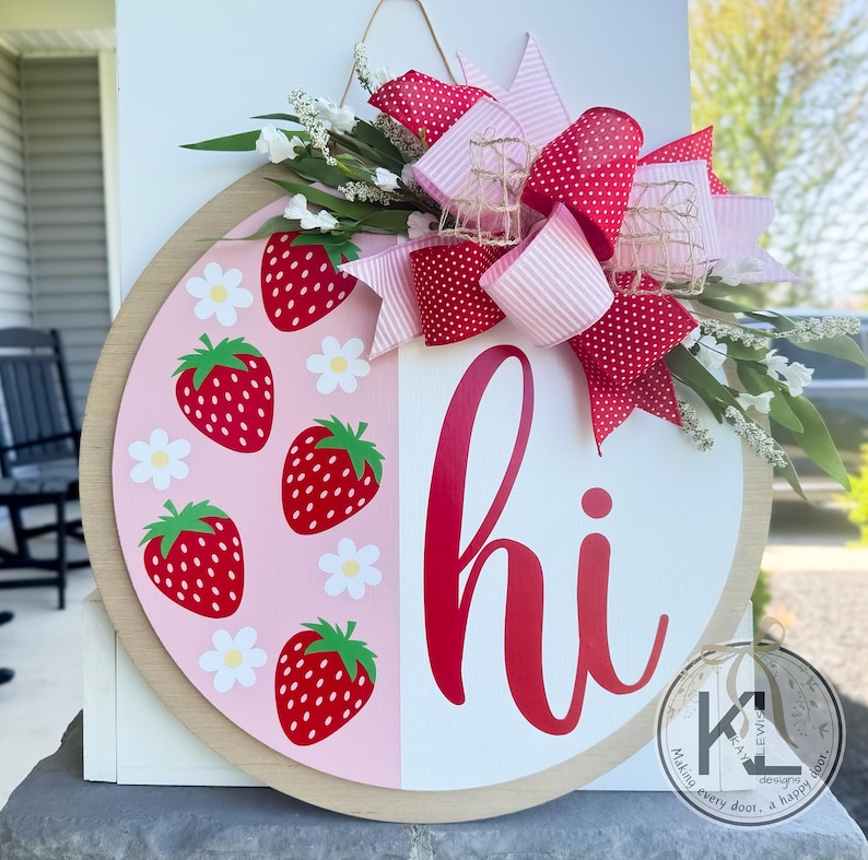 May include: A pink and white round door hanger with a red "hi" and a strawberry pattern. The door hanger is decorated with a pink and red polka dot bow.