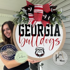 May include: A round wooden sign with the words "GEORGIA Bulldogs" in black and red. The sign features a bulldog logo and is decorated with a bow made of red and white striped ribbon, football-themed ribbon, and greenery. The sign is a genuine college product.