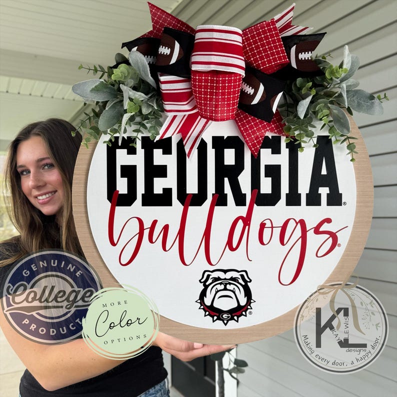 May include: A round wooden sign with the words "GEORGIA Bulldogs" in black and red. The sign features a bulldog logo and is decorated with a bow made of red and white striped ribbon, football-themed ribbon, and greenery. The sign is a genuine college product.