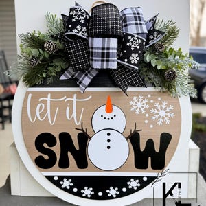 May include: A round wooden sign with the text "let it snow" in black lettering. A snowman is in the center of the sign. The sign is decorated with a black and white plaid bow and greenery.