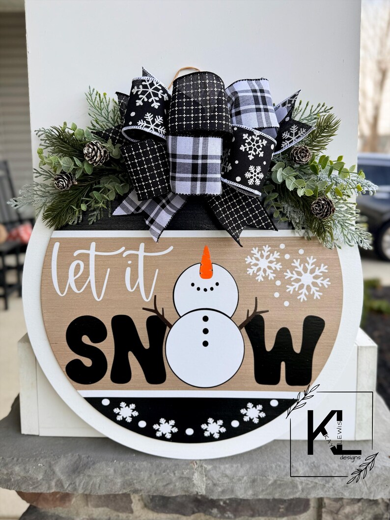 May include: A round wooden sign with the text "let it snow" in black lettering. A snowman is in the center of the sign. The sign is decorated with a black and white plaid bow and greenery.