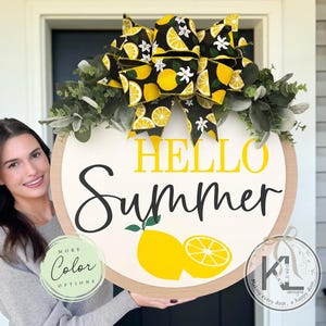 Op de afbeelding: Een rond houten deurbord met de tekst HELLO SUMMER in gele en zwarte letters. Het bord is versierd met een grote strik met een patroon van gele citroenen en witte bloemen, geaccentueerd door groene eucalyptusbladeren en twee citroenafbeeldingen.