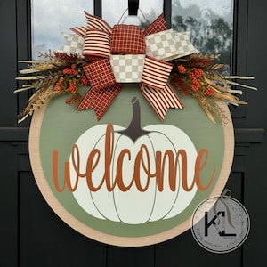 Pode incluir: Uma placa de boas-vindas redonda de madeira com um design de ab&oacute;bora. A placa apresenta uma ab&oacute;bora branca com uma haste cinza e a palavra "welcome" em escrita laranja. &Eacute; decorada com um grande la&ccedil;o e folhagem de outono.