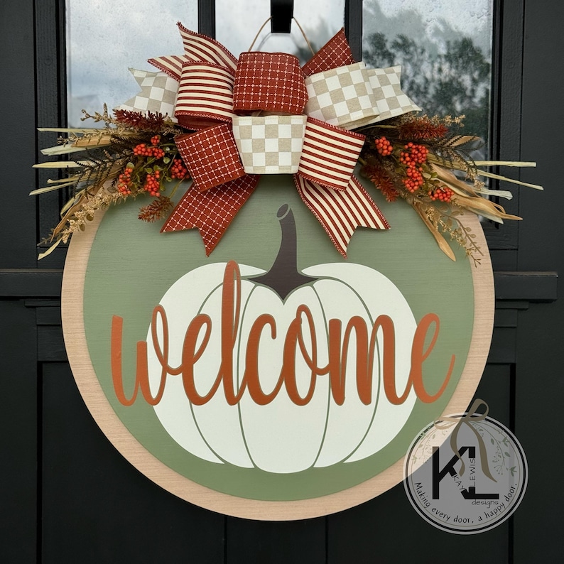Pode incluir: Uma placa de boas-vindas redonda de madeira com um design de ab&oacute;bora. A placa apresenta uma ab&oacute;bora branca com uma haste cinza e a palavra "welcome" em escrita laranja. &Eacute; decorada com um grande la&ccedil;o e folhagem de outono.