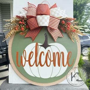Pode incluir: Uma placa de boas-vindas redonda de madeira com fundo verde e um desenho de ab&oacute;bora branca. A palavra "welcome" est&aacute; escrita em laranja. A placa &eacute; decorada com um la&ccedil;o e folhagem de outono.