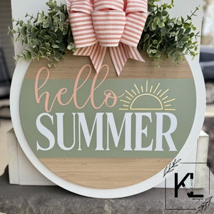 Pu&ograve; includere: Un cartello rotondo in legno con un bordo bianco e uno sfondo verde e legno. Il cartello dice "hello summer" in lettere bianche con una grafica di sole giallo. Il cartello &egrave; decorato con un fiocco a righe rosa e bianco e fogliame verde.