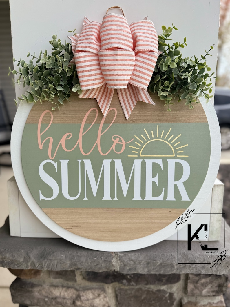 Pu&ograve; includere: Un cartello rotondo in legno con un bordo bianco e uno sfondo verde e legno. Il cartello dice "hello summer" in lettere bianche con una grafica di sole giallo. Il cartello &egrave; decorato con un fiocco a righe rosa e bianco e fogliame verde.