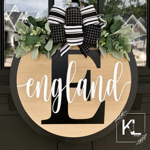 May include: A black and white wreath with a black and white bow and the name "England" in white lettering on a wooden round door hanger.