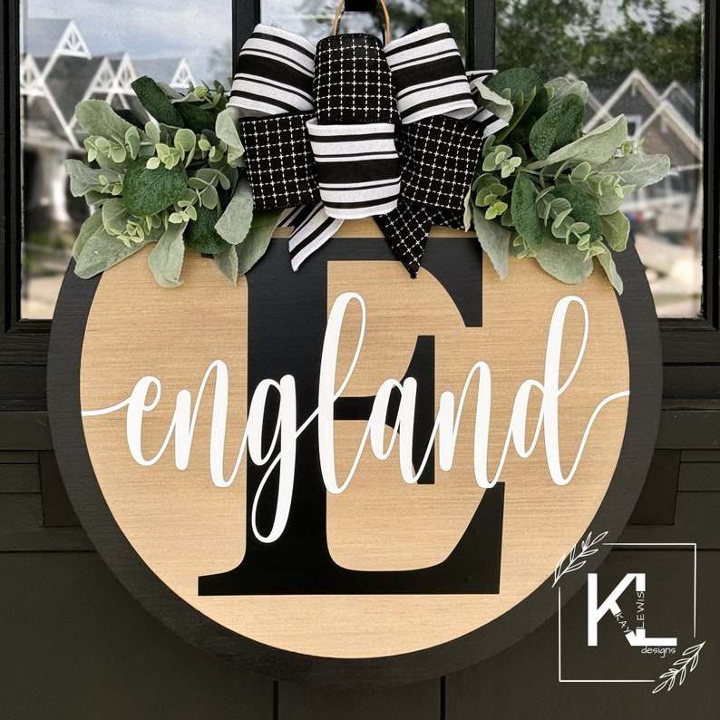 May include: A black and white wreath with a black and white bow and the name "England" in white lettering on a wooden round door hanger.