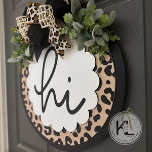 May include: Round wooden door sign with a black rim and a leopard print background. A white scalloped shape in the center says "hi" in black script. The sign is decorated with greenery and a bow with leopard print ribbon.
