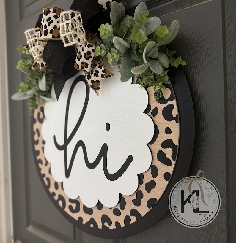 May include: Round wooden door sign with a black rim and a leopard print background. A white scalloped shape in the center says "hi" in black script. The sign is decorated with greenery and a bow with leopard print ribbon.