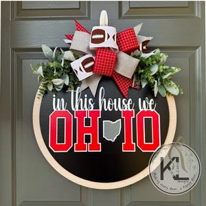May include: A round wooden door sign with a black center and light wood frame. The text reads in this house we OH-IO with a gray map of Ohio replacing the letter I. A decorative bow with football patterns and green foliage sits on top.