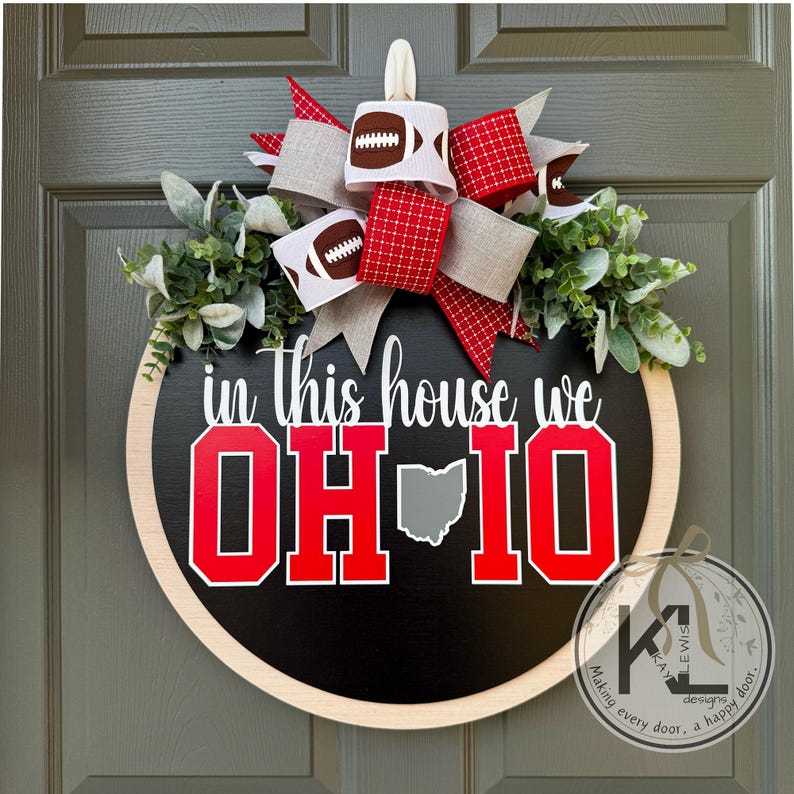 May include: A round wooden door sign with a black center and light wood frame. The text reads in this house we OH-IO with a gray map of Ohio replacing the letter I. A decorative bow with football patterns and green foliage sits on top.
