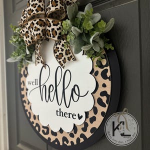 May include: Round wooden welcome sign with a leopard print border and a black frame. The sign features the words "well hello there" in elegant script. A leopard print bow and greenery add a decorative touch.