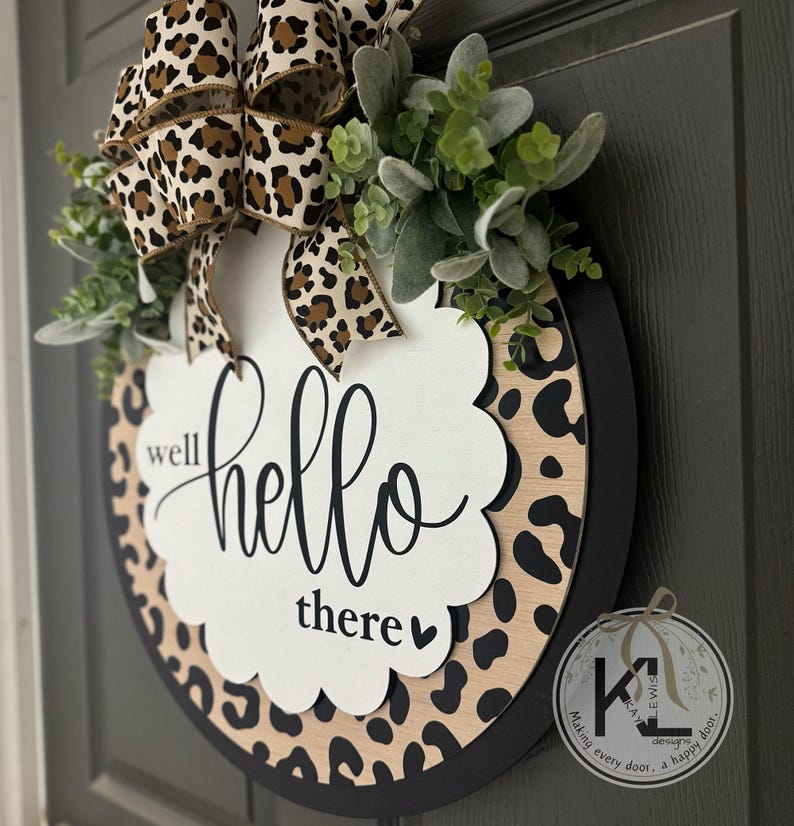 May include: Round wooden welcome sign with a leopard print border and a black frame. The sign features the words "well hello there" in elegant script. A leopard print bow and greenery add a decorative touch.