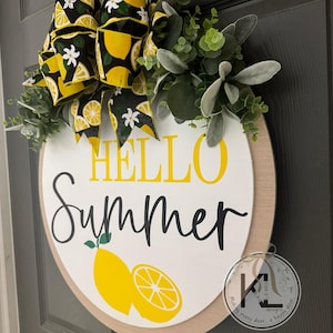 Op de afbeelding: Een rond houten deurbord met de tekst HELLO in het geel en Summer in zwarte schrijfletters. Onder de tekst staan twee gele citroenillustraties. Het bord is versierd met een grote zwarte strik met een patroon van gele citroenen en witte bloemen, samen met groen eucalyptusblad.