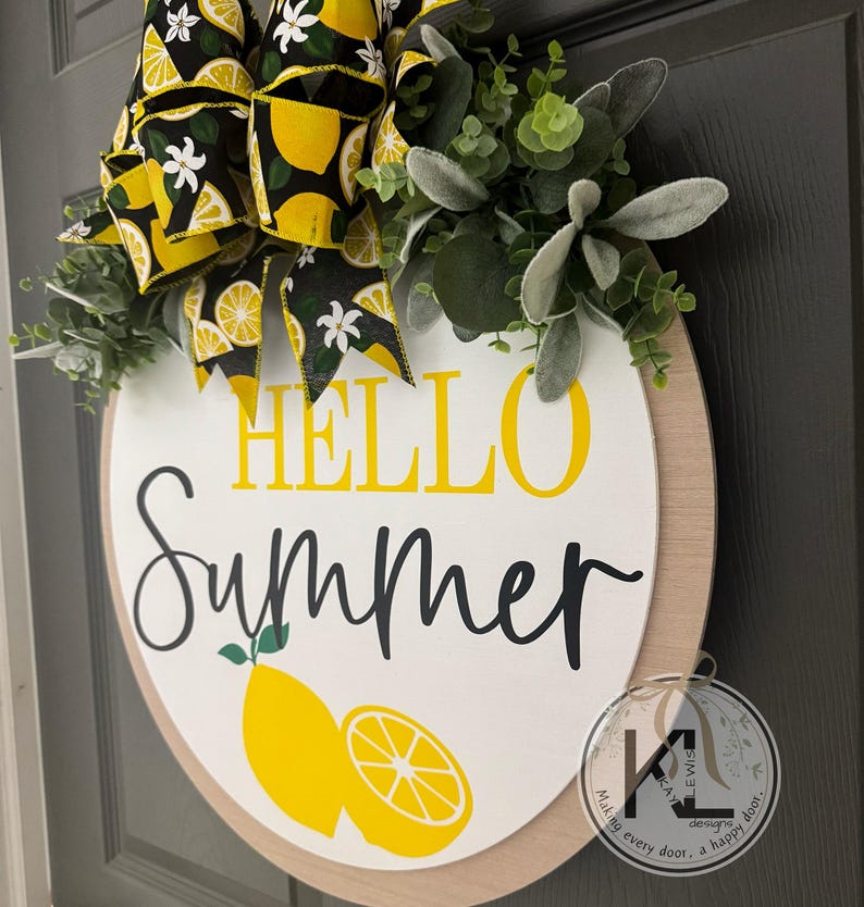 Op de afbeelding: Een rond houten deurbord met de tekst HELLO in het geel en Summer in zwarte schrijfletters. Onder de tekst staan twee gele citroenillustraties. Het bord is versierd met een grote zwarte strik met een patroon van gele citroenen en witte bloemen, samen met groen eucalyptusblad.