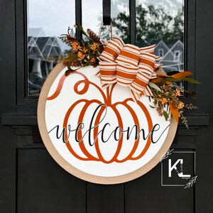May include: A round wooden door hanger with a white background and an orange pumpkin silhouette. The word "welcome" is written in black cursive script inside the pumpkin. The pumpkin is topped with a white and orange striped bow and fall foliage.