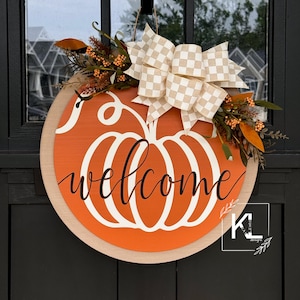 May include: A round wooden door hanger with a pumpkin design and the word "welcome" in black script. The pumpkin is orange with a white outline and is decorated with a beige and white checkered bow and fall foliage.