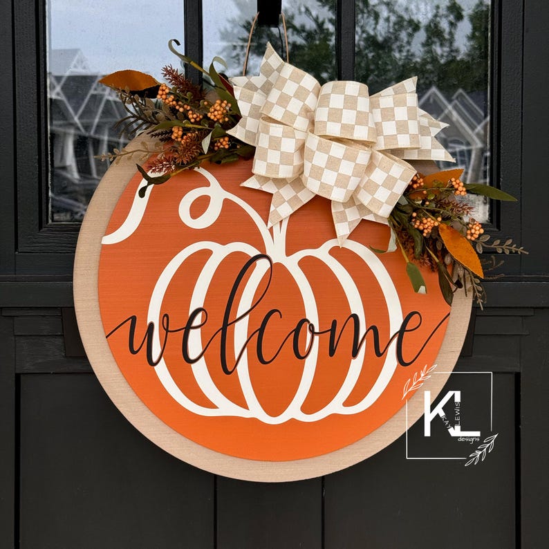 May include: A round wooden door hanger with a pumpkin design and the word "welcome" in black script. The pumpkin is orange with a white outline and is decorated with a beige and white checkered bow and fall foliage.