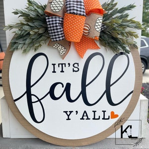 May include: A round wooden sign with the text "It's fall y'all" in black script with a small orange heart. The sign is decorated with a fall-themed bow made of orange and black checkered ribbon, orange polka dot ribbon, and burlap ribbon with a pumpkin design.