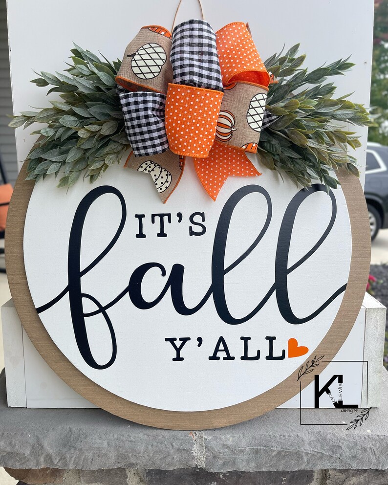May include: A round wooden sign with the text "It's fall y'all" in black script with a small orange heart. The sign is decorated with a fall-themed bow made of orange and black checkered ribbon, orange polka dot ribbon, and burlap ribbon with a pumpkin design.