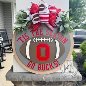 May include: A circular wooden door sign with a football theme. It features a football design with the number "0" in red, the text "'TIS THE SEASON GO BUCKS!" and a decorative bow with football accents. Greenery adds a natural touch.