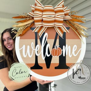 May include: A round wooden door sign featuring a large black letter M, the white script text welcome, and a small orange pumpkin graphic. The sign has horizontal stripes in shades of orange and tan, topped with a large orange and white striped bow.