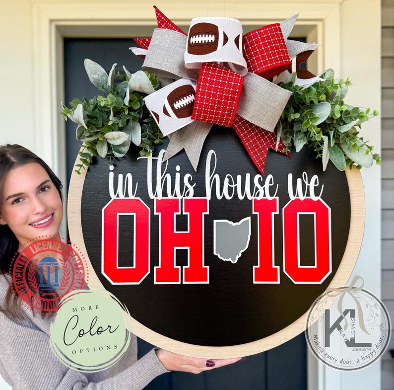 May include: A round wooden door sign with a black background featuring the white text "in this house we" above large red block letters "OH" and "IO" with a gray outline of the state of Ohio in between. A large bow with football and red patterns sits on top with green foliage.